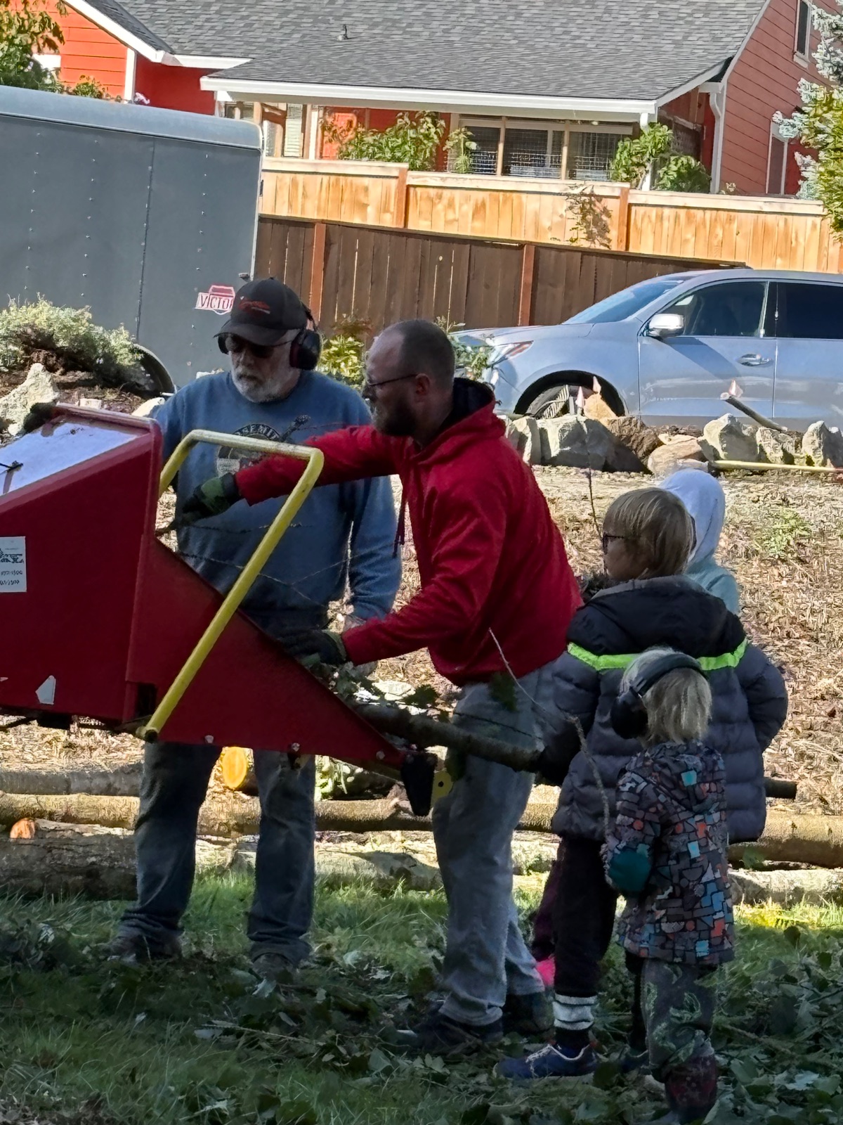 Volunteers feeding cut branches into a wood chipper while children in ear protection look on